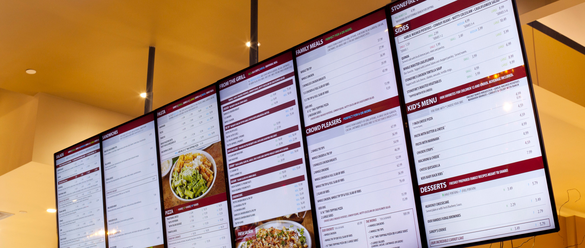 Six vertical digital menu boards are hung from a restaurant ceiling.