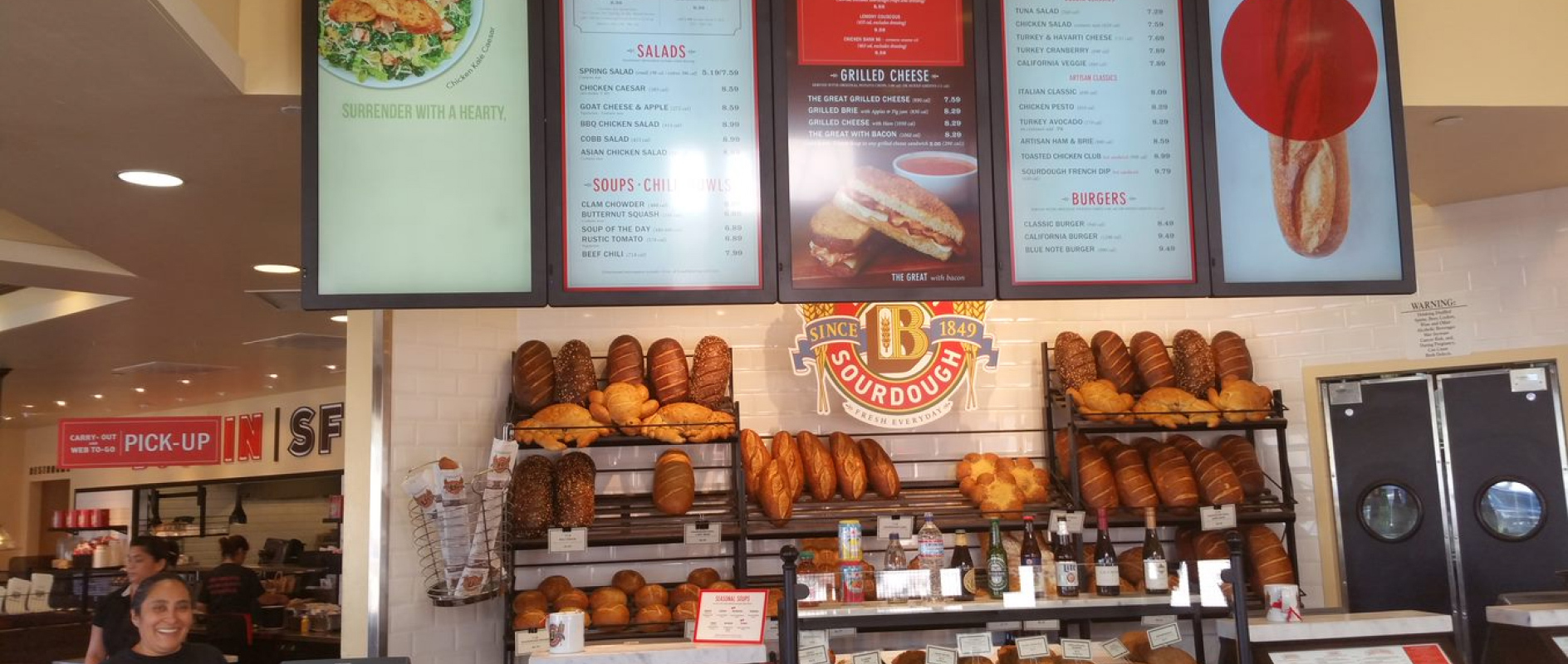 A tall rack of baked bread loaves underneath five vertical video menu screens showing soups and sandwiches.