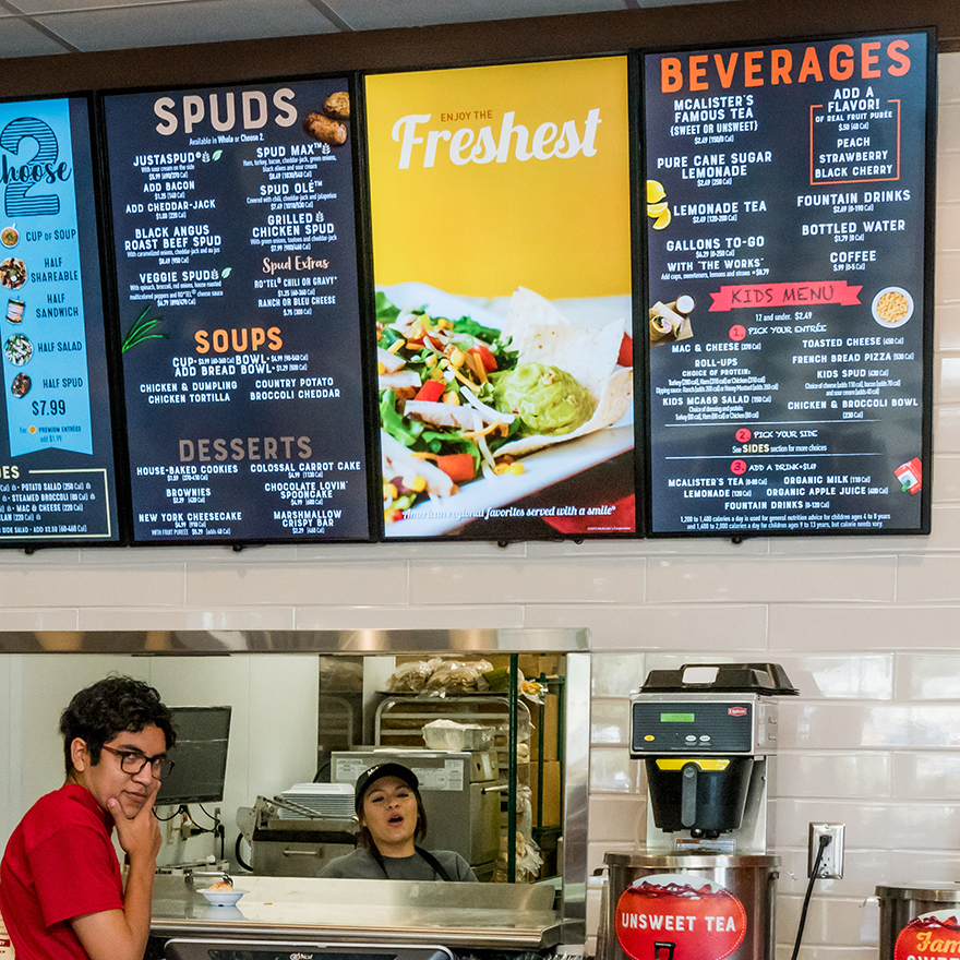 Two people are placing an order with a cashier in front a row of eight vertical video boards advertising the restaurant's menu.