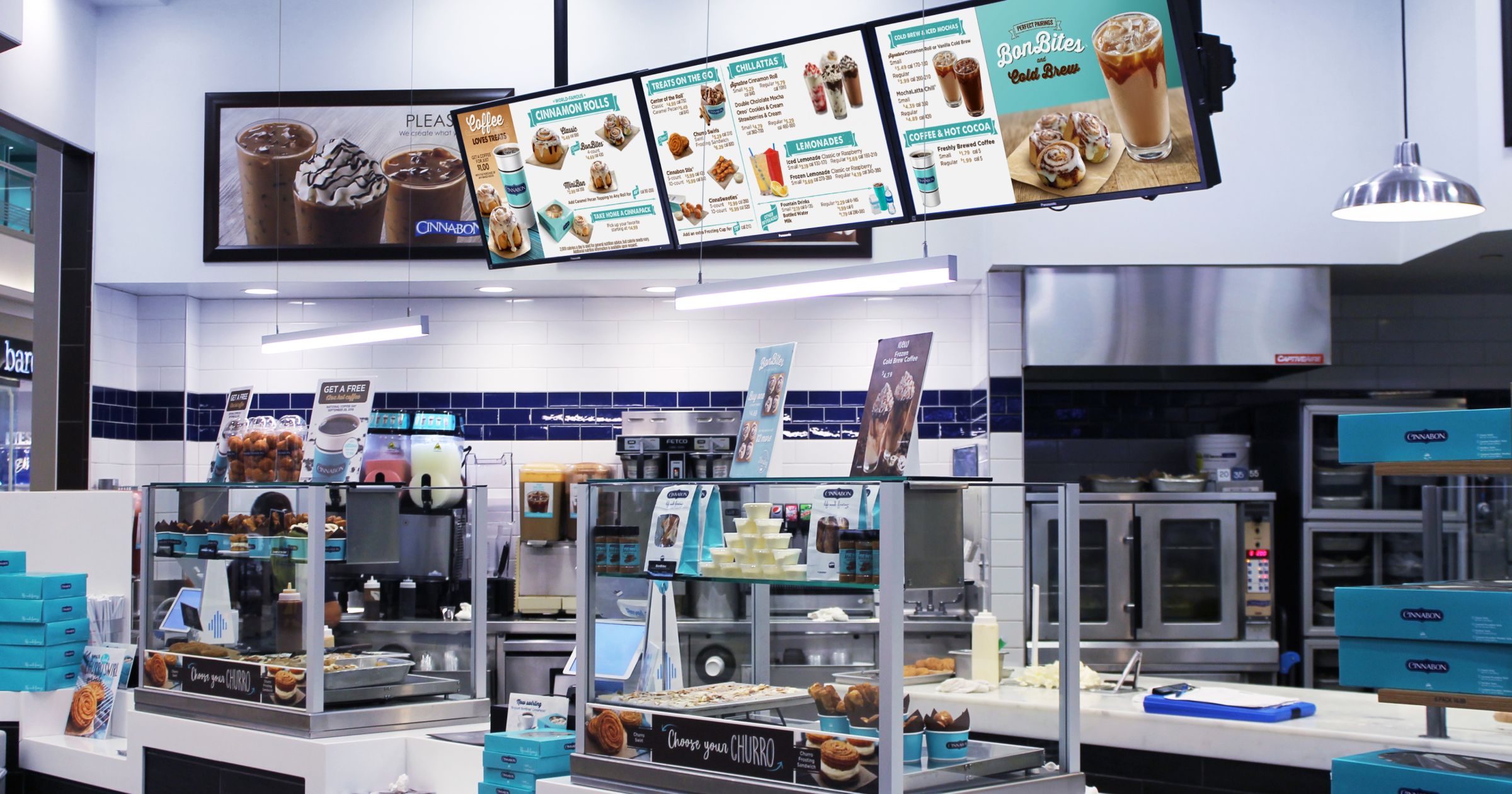 A brightly lit Cinnabon storefront showing white countertops, two glass display cases, and three digital menus hung overhead. 